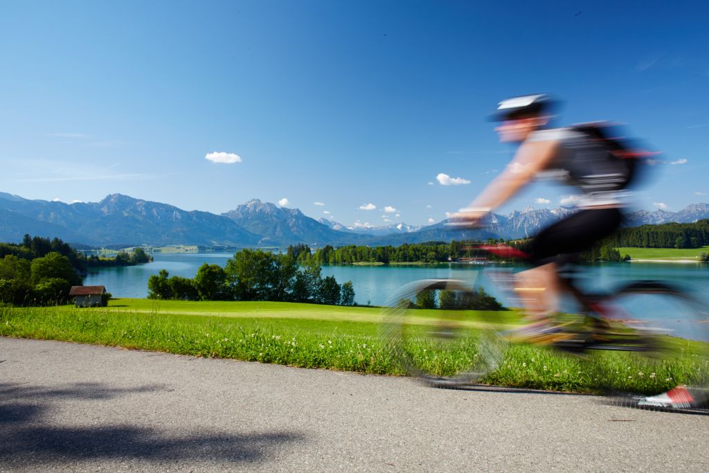 Ein verschwommener Radfahrer fährt an einem malerischen Seeufer mit grünem Gras, Bäumen, ruhigem Wasser und fernen Bergen unter einem blauen Himmel mit vereinzelten Wolken vorbei.