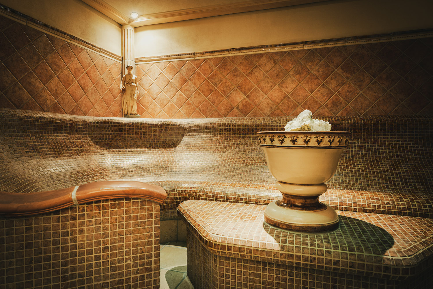 Ein luxuriöses Dampfbad mit Bänken aus braunen Mosaikfliesen, einer dekorativen Urne mit weißen Handtüchern und warmer Umgebungsbeleuchtung. Die Wände sind mit einem Kachelmuster versehen und in der Ecke steht eine kleine Statue.