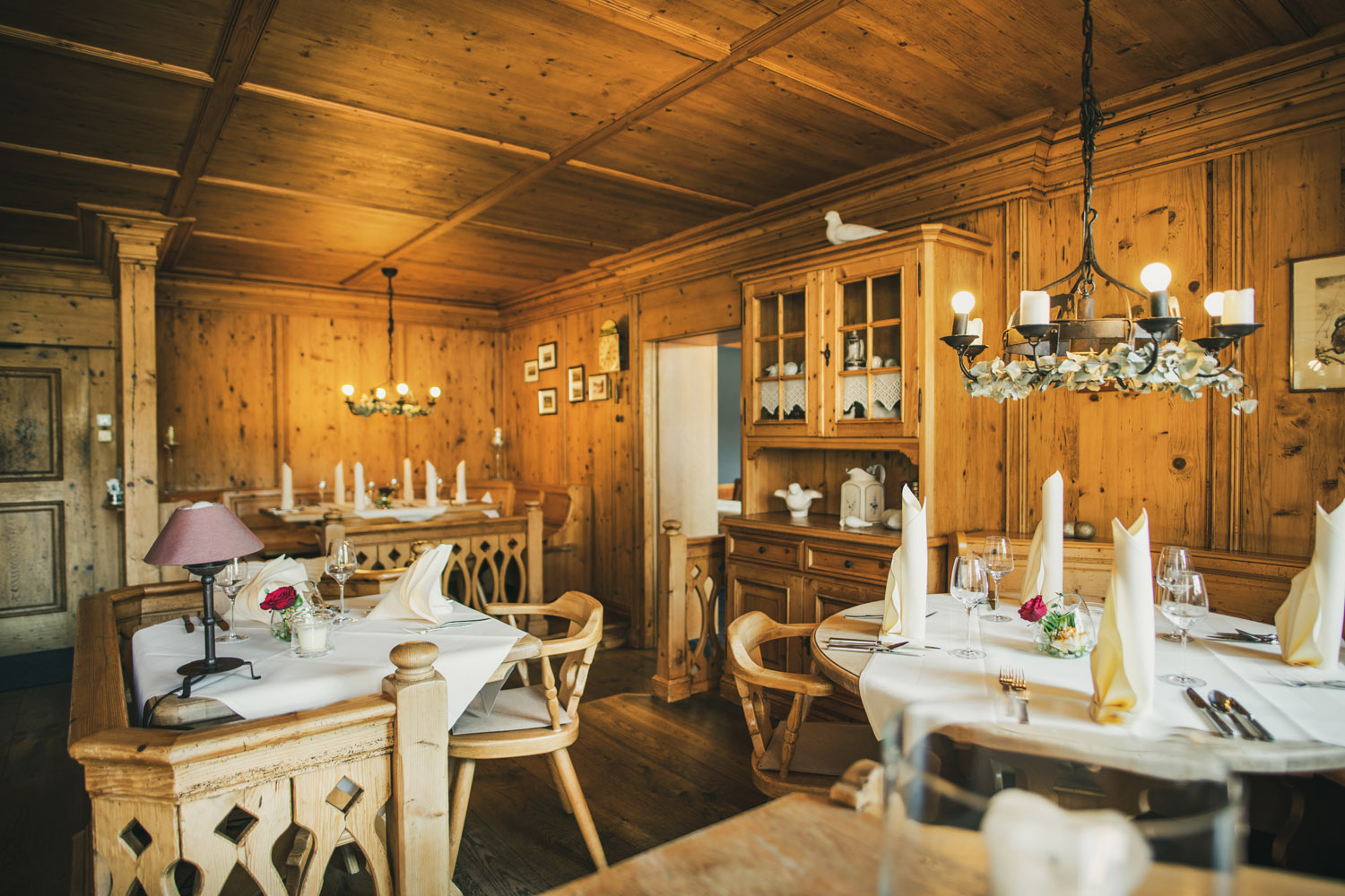 Ein gemütlicher Speisesaal mit hölzernen Wänden und Decken, ordentlich gedeckten Tischen mit weißen Tüchern, gefalteten Servietten, Weingläsern und Blumenkränzen, sanft beleuchtet von Kronleuchtern und natürlichem Licht.