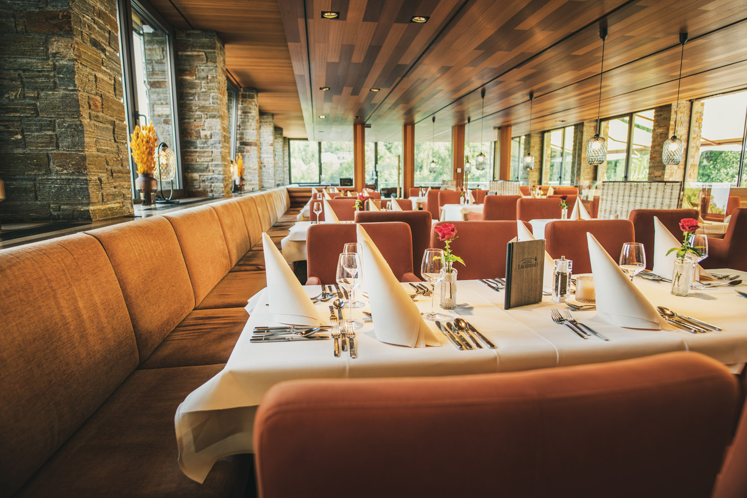 Elegantes Restaurantinterieur mit ordentlich gedeckten Tischen, gefalteten weißen Servietten, Glaswaren, Besteck und rosa Rosen in Vasen. Die warme Beleuchtung, die Holzdecke und die großen Fenster schaffen eine gemütliche, einladende Atmosphäre.