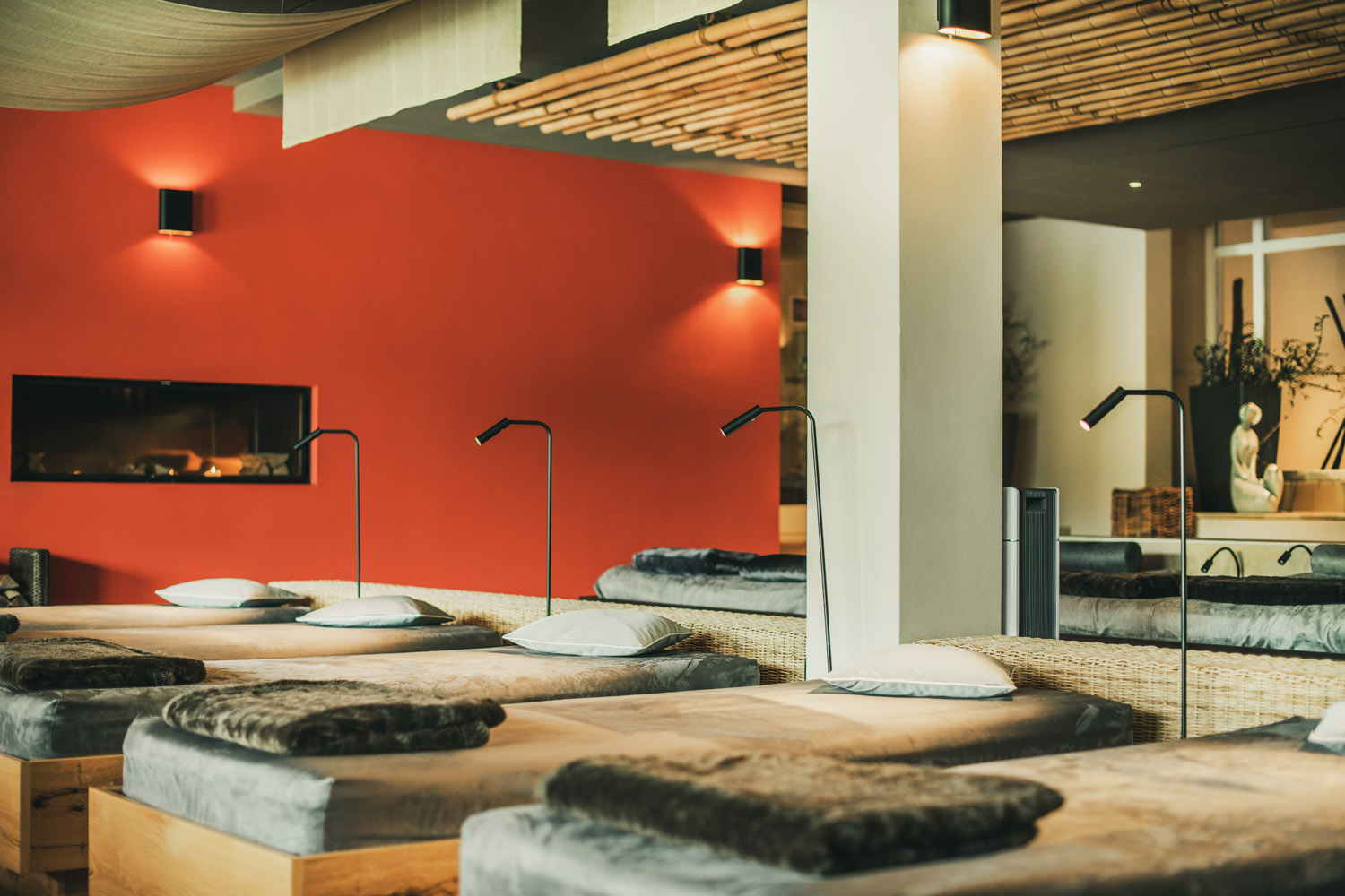 Eine moderne Entspannungslounge mit mehreren aneinandergereihten Einzelbetten, jedes mit grauen Decken, Kissen und einzelnen Leselampen. Der Raum verfügt über eine rote Akzentwand, Bambuselemente und eine sanfte Raumbeleuchtung.