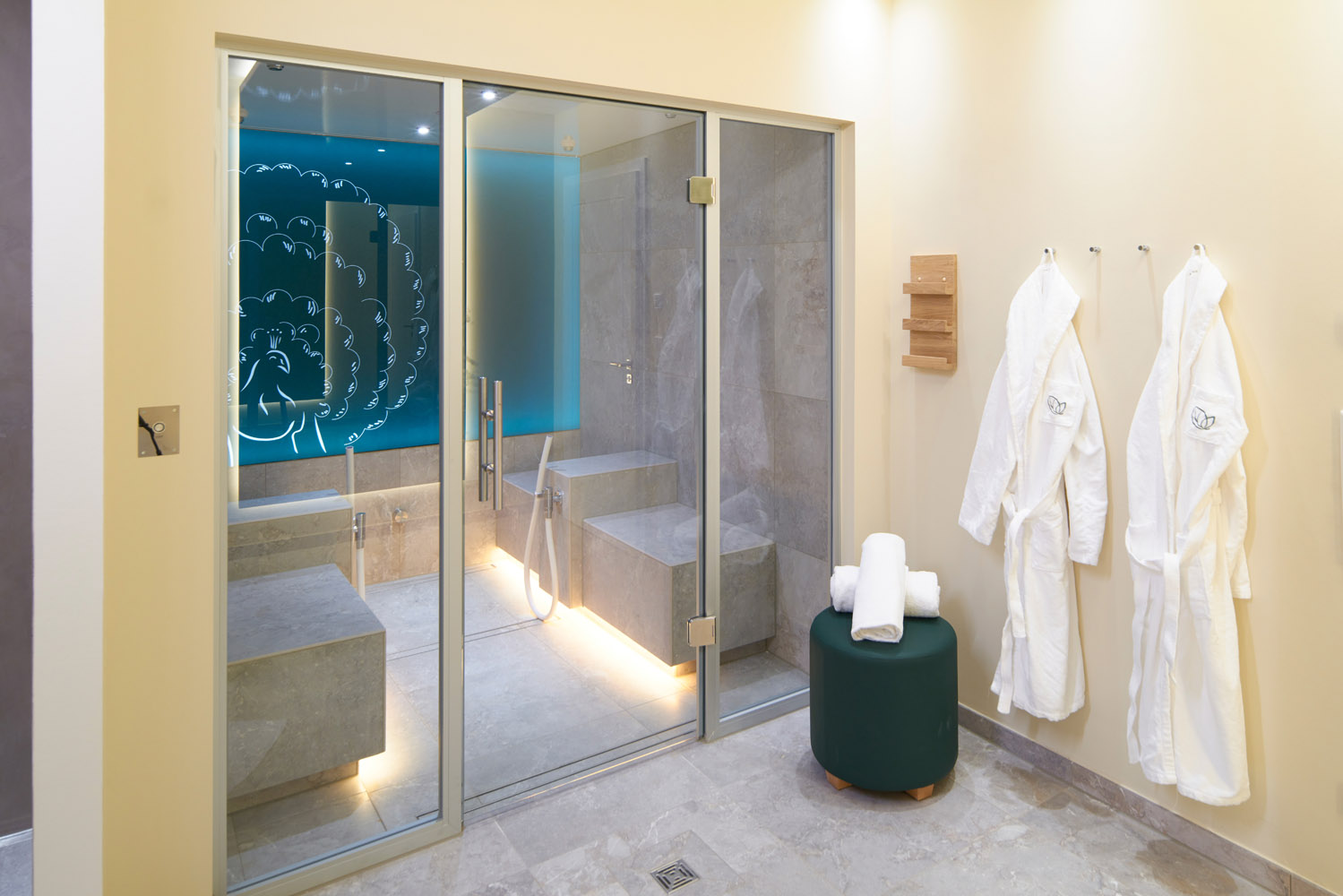 Ein moderner Spa-Raum mit einer gläsernen Dampfkabine, blau beleuchteter Wandkunst, zwei weißen Bademänteln, die an Haken hängen, einem grünen Hocker mit gerollten Handtüchern und beigen Wänden und Böden.