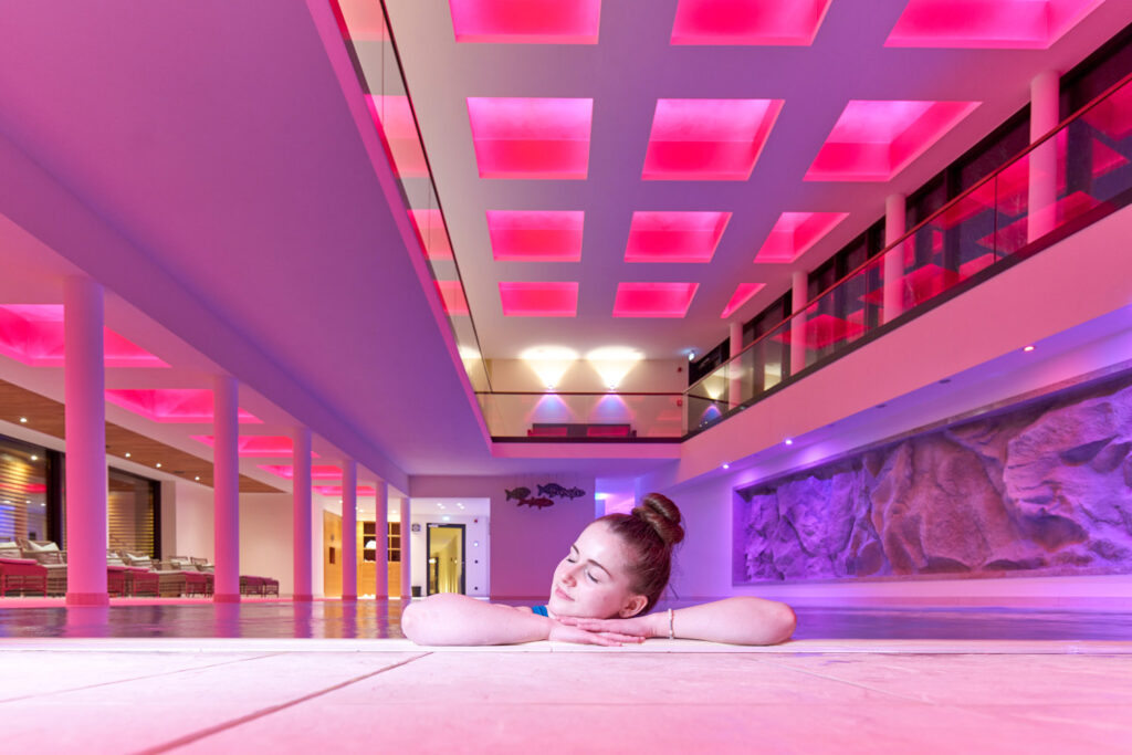 Eine Frau mit einem Dutt im Haar stützt ihr Kinn auf die Hände, die auf dem Boden eines modernen, geräumigen Raums mit rosa und lila Deckenleuchten und einer großen dekorativen Wand liegen.