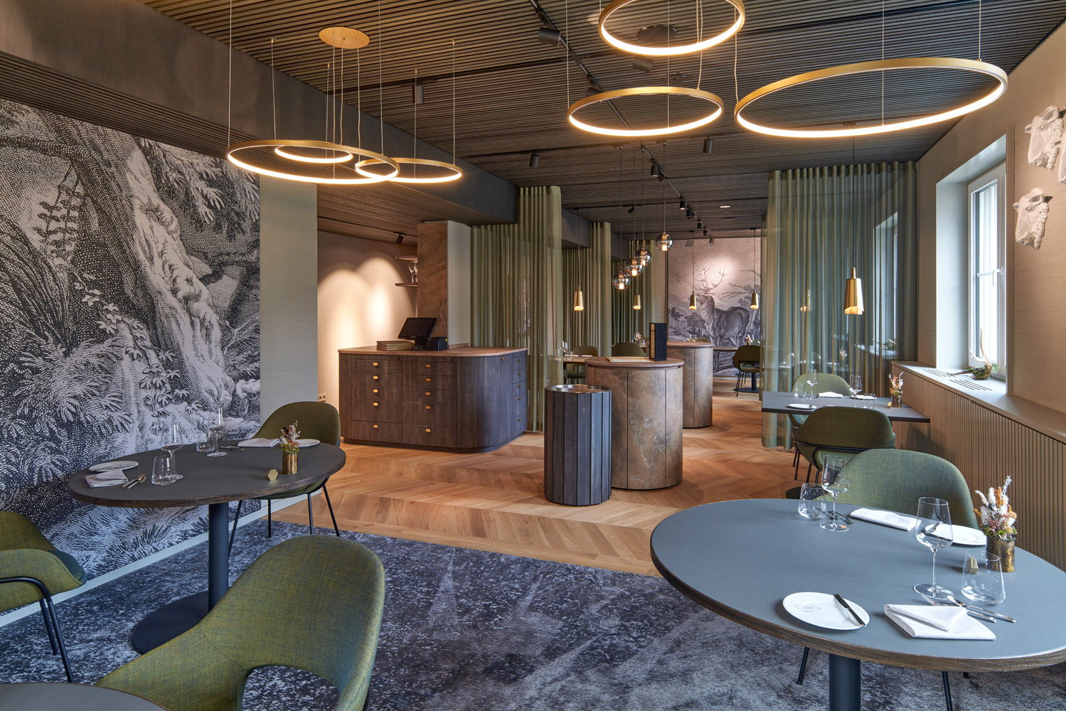 Modernes Restaurantinterieur mit runden Hängelampen, grünen Stühlen, runden Tischen und Holzakzenten. Ein künstlerisches schwarz-weißes Wandgemälde und sanftes natürliches Licht aus mehreren Fenstern schaffen eine stilvolle, einladende Atmosphäre.