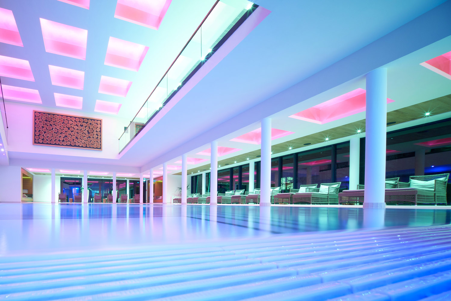 Moderner Innenpoolbereich mit Säulen, Liegestühlen und einer Decke mit leuchtenden rosa und lila Lichtern. Große Fenster säumen eine Wand, und das moderne Dekor schafft eine elegante, gehobene Atmosphäre.