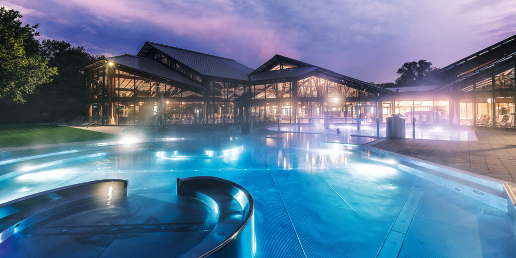 Beleuchtetes Außenbecken mit geschwungenen Metallelementen in der Dämmerung, neben einem modernen Gebäude aus Glas und Holz. Im Mineral-Thermalbad Keidel spiegeln sich sanfte Lichter auf dem Wasser und schaffen eine ruhige Atmosphäre vor einem lila-blauen Himmel.