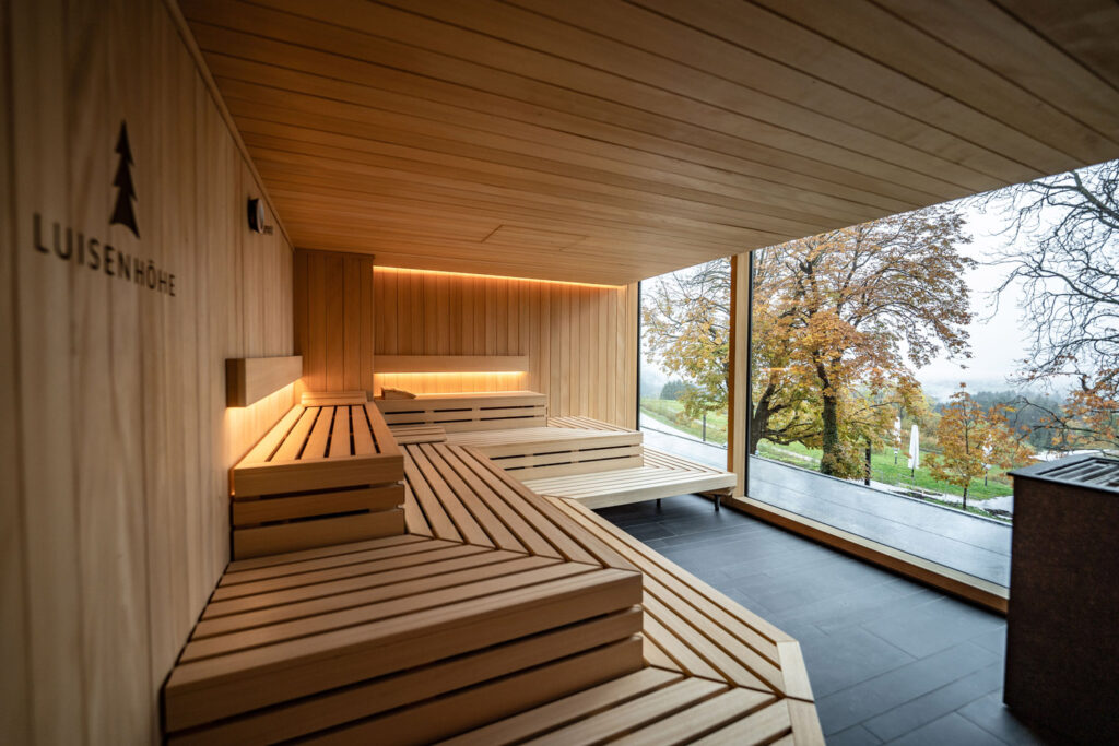 Eine moderne Holzsauna mit Stufenbänken, einem großen, bodentiefen Fenster, das den Blick auf Bäume mit Herbstlaub freigibt, und einer warmen Raumbeleuchtung. Das Wort LUISENHÖHE und ein Baumlogo sind an der Wand angebracht.