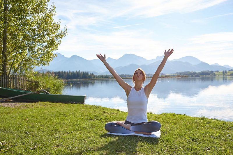 Eine Frau sitzt im Schneidersitz auf einer Matte im Freien an einem See und hebt ihre Arme in den Himmel. Umgeben von Bergen und Bäumen, umarmt sie Mental Wellness fürs Wohlbefinden in dieser friedlichen, natürlichen Umgebung unter blauem Himmel und Wolken.