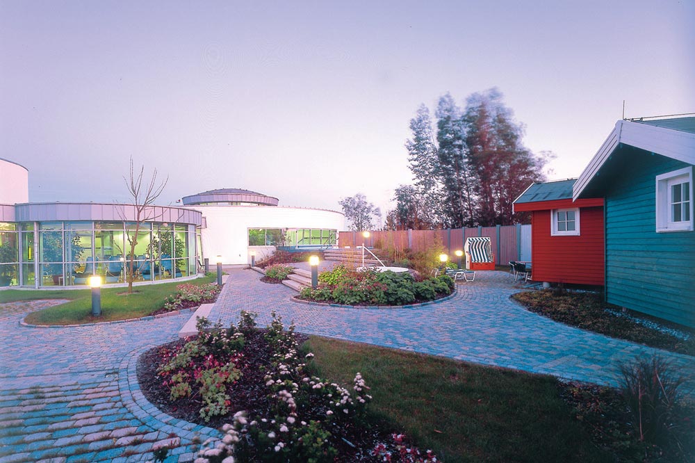 Ein landschaftlich gestalteter Innenhof mit geschwungenen Wegen, Gärten und modernen Rundbauten mit großen Fenstern auf der linken Seite. Auf der rechten Seite stehen bunte kleine Holzhäuser und Terrassenmöbel. Der Himmel ist klar und die Bäume sind im Hintergrund.