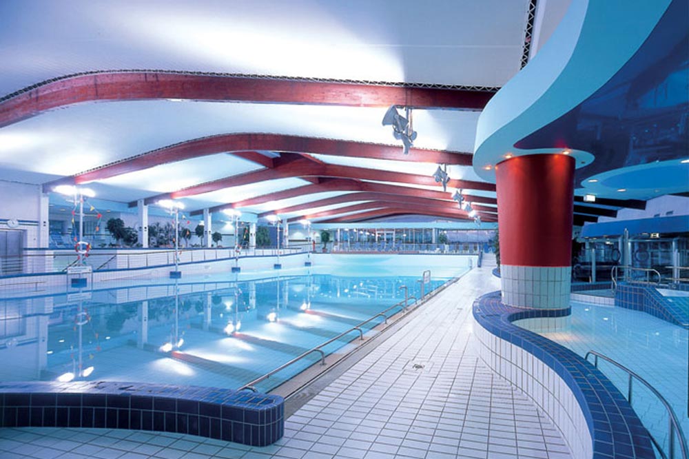 Hallenbad mit sauberem, blauem Wasser, weiß gefliestem Boden, roten Deckenbalken und geschwungener, moderner Architektur. Der Bereich ist gut beleuchtet, leer und verfügt über Handläufe und Stufen, die in den Pool führen.