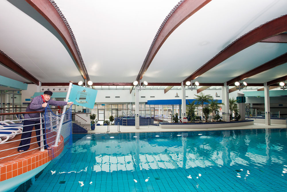 Eine Person in Uniform steht am Geländer eines Hallenbades und schaut ins Wasser. Der Schwimmbadbereich ist hell erleuchtet, mit großen Deckenbalken versehen und mit Pflanzen dekoriert. Die Atmosphäre ist sauber und modern.