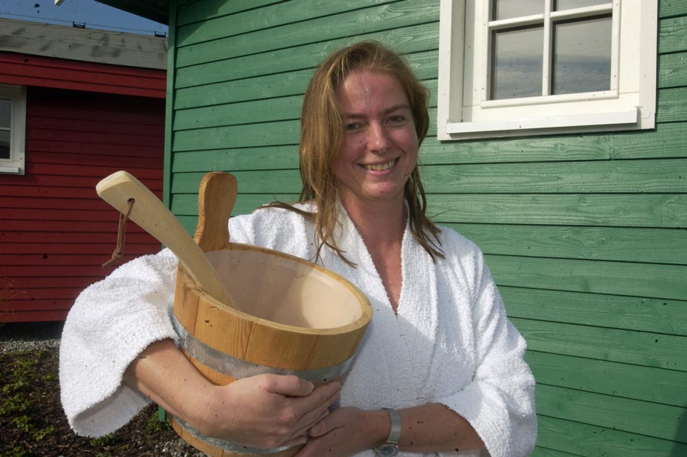 Eine lächelnde Frau in einem weißen Bademantel steht draußen an einem grünen Holzgebäude und hält einen hölzernen Saunakübel und eine Kelle in der Hand.