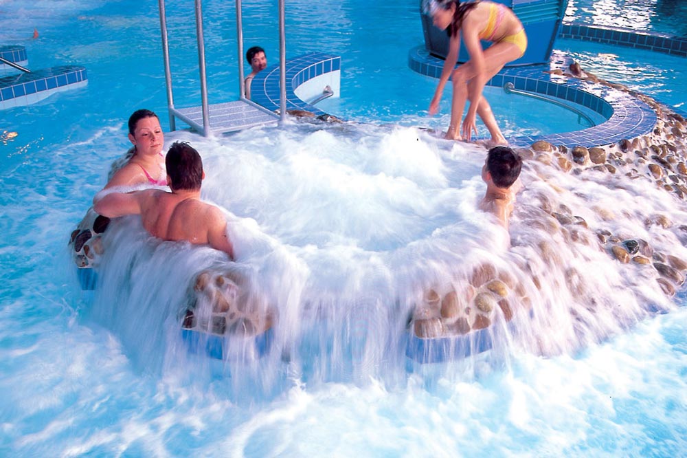 Mehrere Personen entspannen und spielen in einem Hallenbad mit sprudelndem Wasser und einem runden Whirlpool aus Stein, aus dem Wasser in das Becken fließt. Ein Kind steht am Rand, andere genießen das warme Wasser.