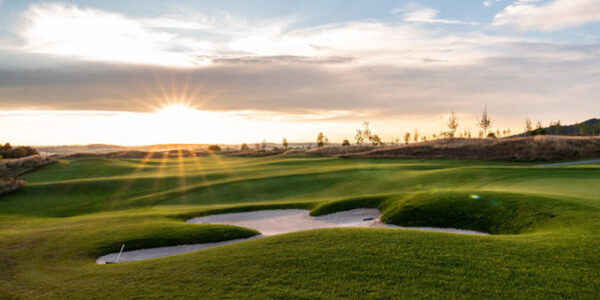 Sonnenuntergang über einem Golfplatz mit ordentlich getrimmtem grünen Gras, Sandbunker im Vordergrund und ein paar Bäumen, die sich gegen den teilweise bewölkten Himmel abzeichnen - perfekt für einen entspannenden Wander-Wellnessurlaub in der Ortenau.