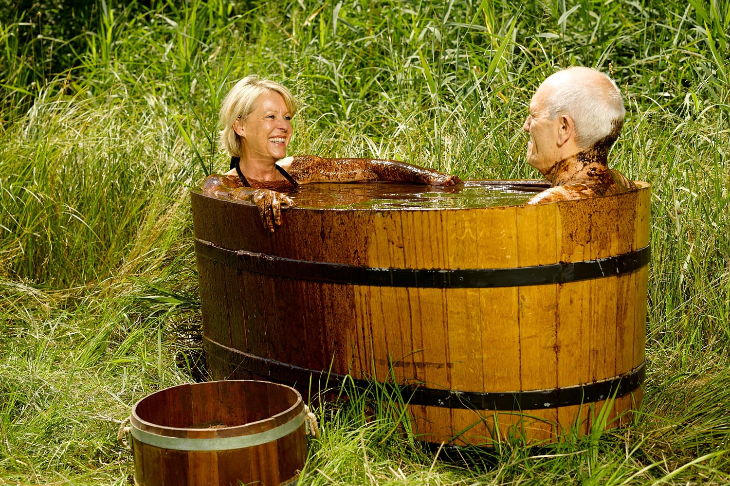 Zwei ältere Erwachsene entspannen sich und lächeln sich an, während sie ein Moorbad in einer großen Holzwanne genießen, die von hohem Gras und Grün umgeben ist - ein typisches Erlebnis in Thermen und Gesundheitsresorts. Eine leere kleinere Wanne steht daneben.