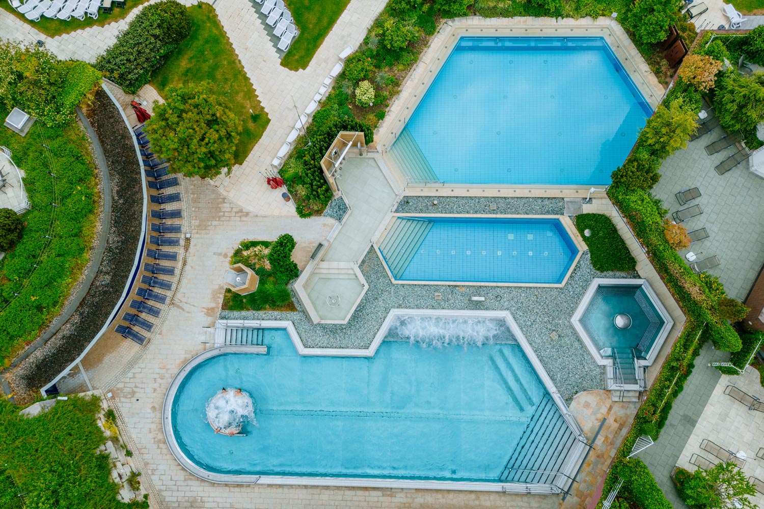 Luftaufnahme eines Freibadkomplexes mit drei verschiedenen Becken, umliegenden Grünflächen, Liegestühlen, Wegen und Landschaftsbereichen. Die Pools variieren in Form und Größe, einschließlich sechseckiger und rechteckiger Designs.