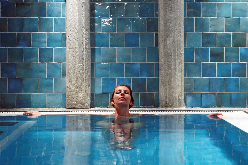 Eine Person entspannt sich in einem Swimmingpool, wobei sie ihre Arme auf den Rand stützt, mit blau gekachelten Wänden im Hintergrund.