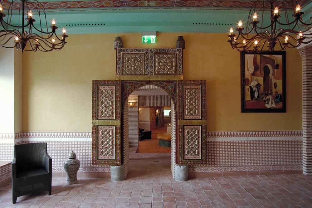 Eine reich verzierte Tür mit verschlungenen Mustern führt in einen anderen Raum. Die Wände sind mit geometrischen Fliesen verziert, von der Decke hängen Kronleuchter, und ein gerahmtes Kunstwerk und eine Keramikvase schmücken den Raum.