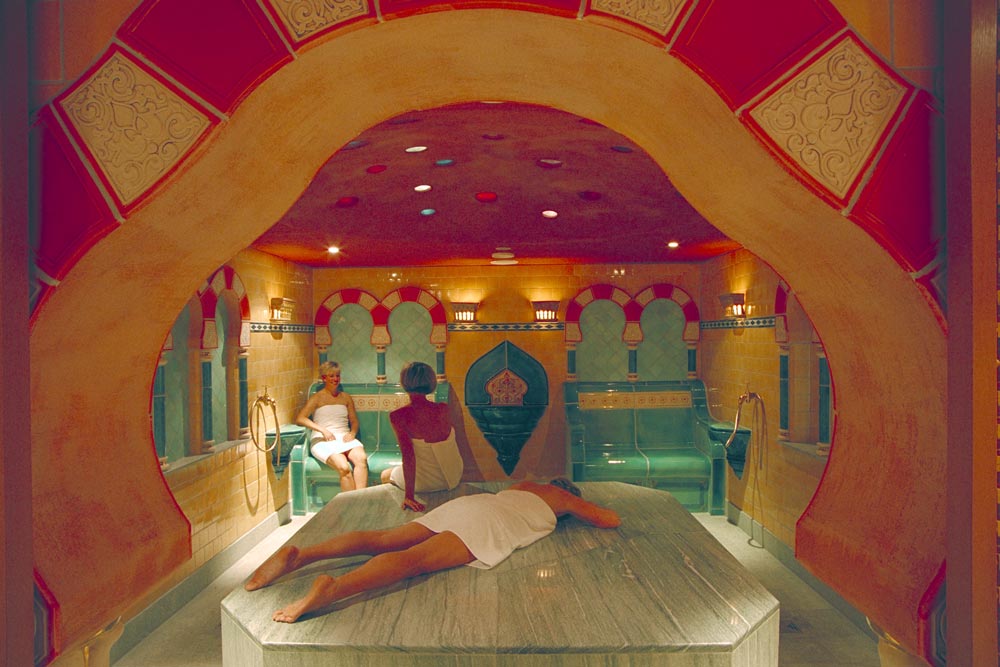 Zwei Personen entspannen sich in einem verschnörkelten Wellness-Raum mit warmer Beleuchtung, gefliesten Wänden und einer gewölbten Decke. Eine Person liegt mit dem Gesicht nach unten auf einer Steinmassageliege, während die andere auf einer Bank im Hintergrund sitzt.