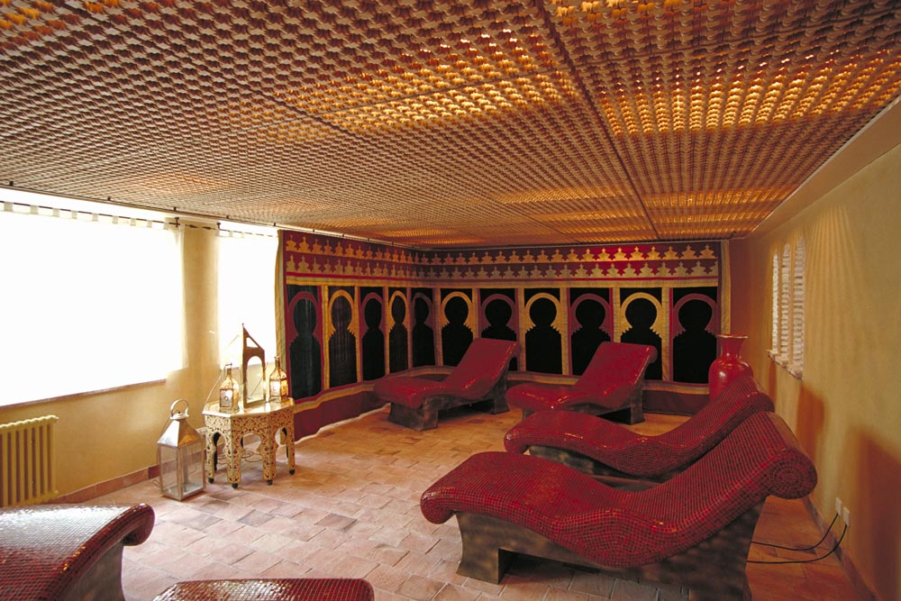 Ein luxuriöser Spa-Ruheraum mit roten Mosaik-Loungesesseln, verzierten Laternen, gemusterten Deckenfliesen und einer kunstvollen Wand mit schwarzen Gewölben und farbigen Akzenten schafft eine exotische, heitere Atmosphäre.