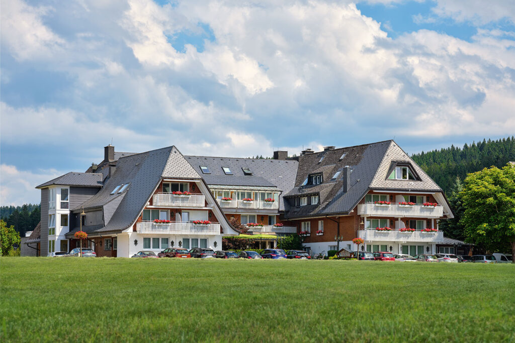 Ein großes Hotelgebäude mit zahlreichen Balkonen und Blumenkästen, das hinter einer grünen Wiese mit geparkten Autos liegt.