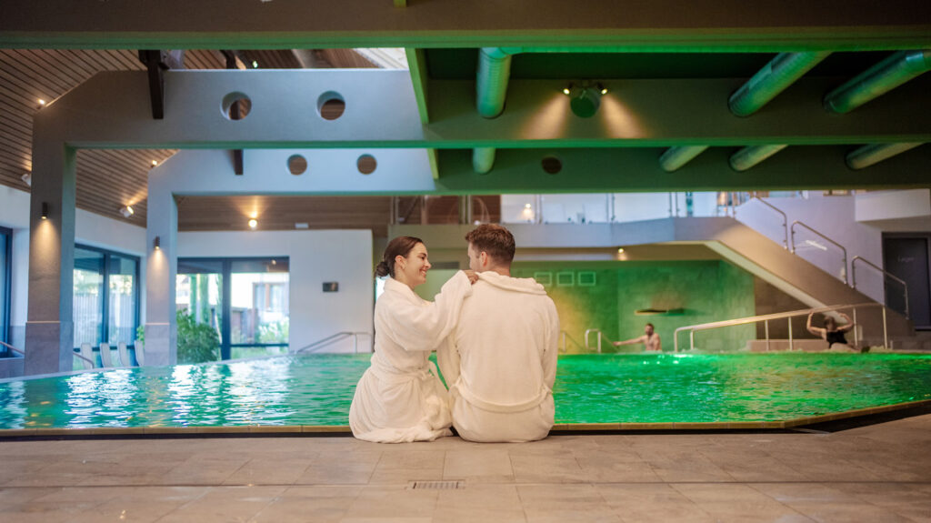 Ein Paar in weißen Bademänteln sitzt an einem Hallenbad und lächelt sich an. Der Pool leuchtet grün unter der Deckenbeleuchtung, und ein paar Leute entspannen sich im Hintergrund. Die Umgebung ist modern und ruhig.
