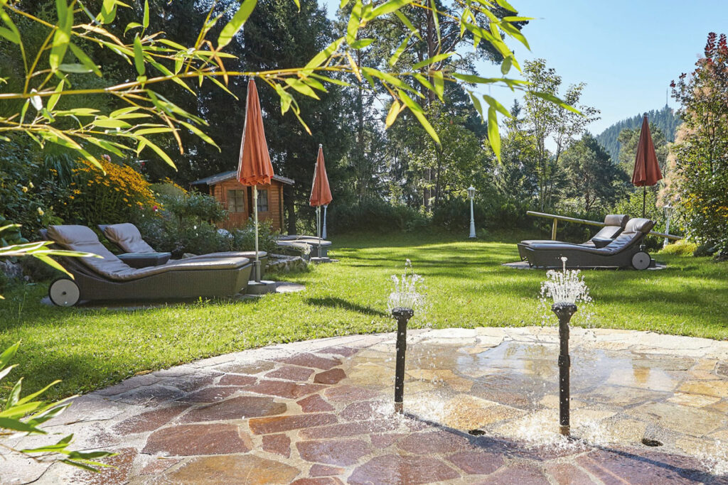 Rasenfläche mit Liegestühlen und geschlossenen orangefarbenen Sonnenschirmen, umgeben von Bäumen und Blumen. Im Vordergrund spritzen Wasserfontänen aus einem gepflasterten Bereich bei hellem Tageslicht.