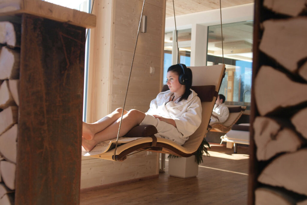 Eine Person in einem weißen Bademantel entspannt sich auf einem Hängesessel in einem Innenraum, trägt Kopfhörer und schaut aus einem großen Fenster. Sonnenlicht strömt herein und schafft eine warme und friedliche Atmosphäre.