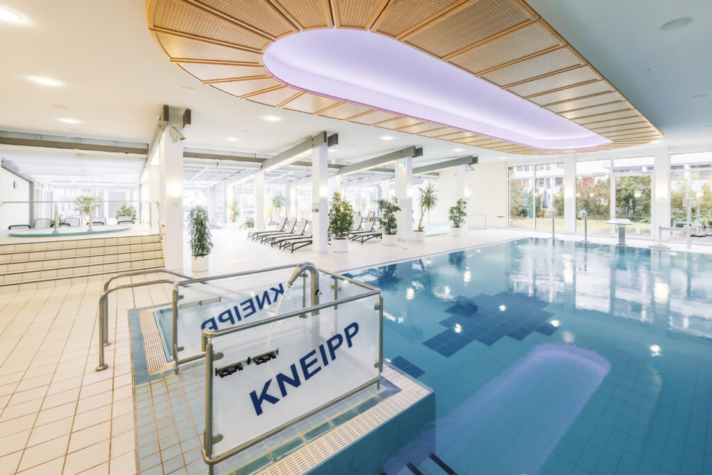 Modernes Hallenbad mit Kneippanlage, Liegestühlen, gefliestem Boden, großen Fenstern und Grünpflanzen. An der Decke befindet sich eine Holzplatte mit violetter Beleuchtung.