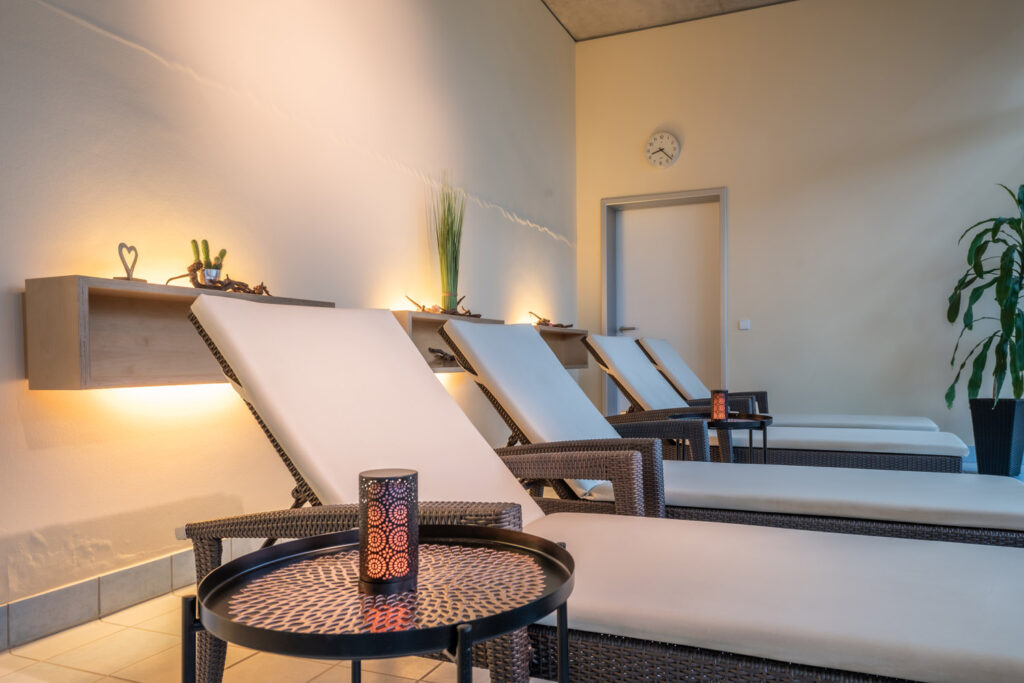 Eine Reihe von gepolsterten Lounge-Sesseln mit Beistelltischen in einem entspannenden, sanft beleuchteten Wellness-Raum, dekoriert mit Kerzen, Pflanzen und minimalistischem Dekor. Eine Wanduhr hängt über einer geschlossenen Tür im Hintergrund.