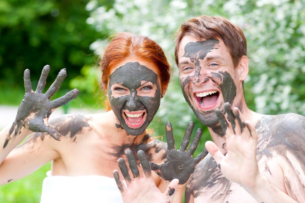 Zwei Menschen lächeln und winken in die Kamera, ihre Gesichter und Hände sind mit schwarzem Schlamm bedeckt. Sie genießen einen spielerischen Moment im Freien inmitten von grünem Laub - eine lustige Wellness für Frauen Aktivität in der Natur.