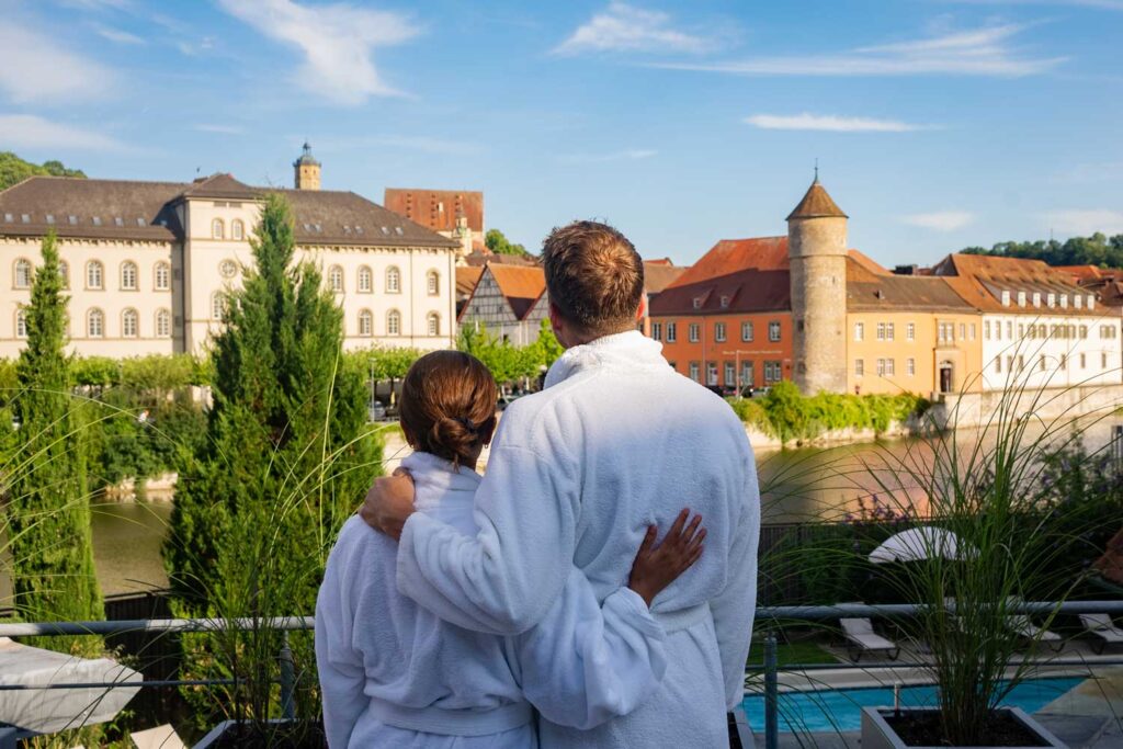 Ein Paar in weißen Bademänteln steht auf einem Balkon, umarmt sich und blickt an einem sonnigen Tag auf eine Stadt am Fluss mit historischen Gebäuden, Bäumen und einem Turm.