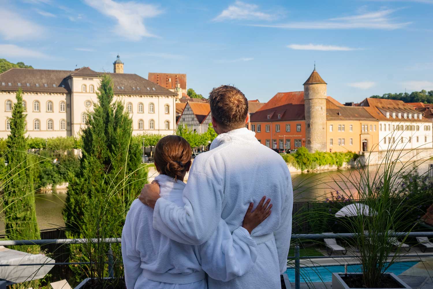 Ein Paar in weißen Bademänteln steht auf einem Balkon, umarmt sich und blickt an einem sonnigen Tag auf eine Stadt am Fluss mit historischen Gebäuden, Bäumen und einem Turm.