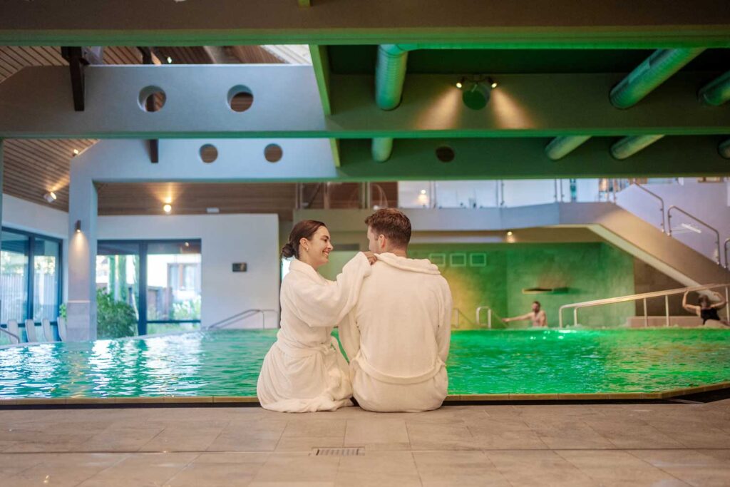 Ein Paar in weißen Bademänteln sitzt an einem Hallenbad und lächelt sich an. Der Pool leuchtet grün unter der Deckenbeleuchtung, und ein paar Leute entspannen sich im Hintergrund. Die Umgebung ist modern und ruhig.
