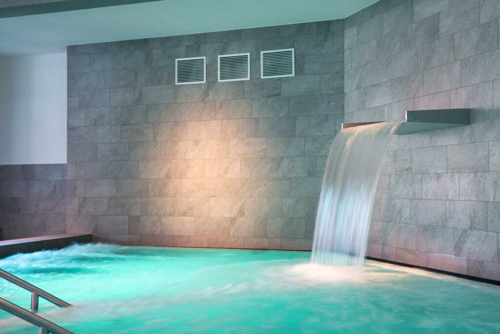 Ein modernes Hallenbad mit türkisfarbenem Wasser verfügt über einen breiten Wasserfall, der von einer grau gefliesten Wand herabfällt; Metallgeländer führen in den Pool, und eine sanfte Beleuchtung schafft eine entspannende Atmosphäre.