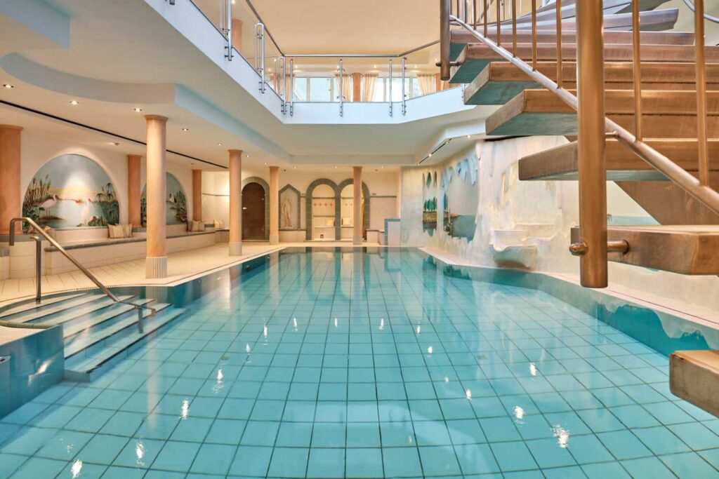 Hallenbad mit klaren blauen Fliesen, umgeben von Säulen, einer Treppe mit Holzgeländer und Wandmalereien mit Naturszenen an den Wänden. Es gibt einen Balkon und einen Sitzbereich oberhalb des Pools.