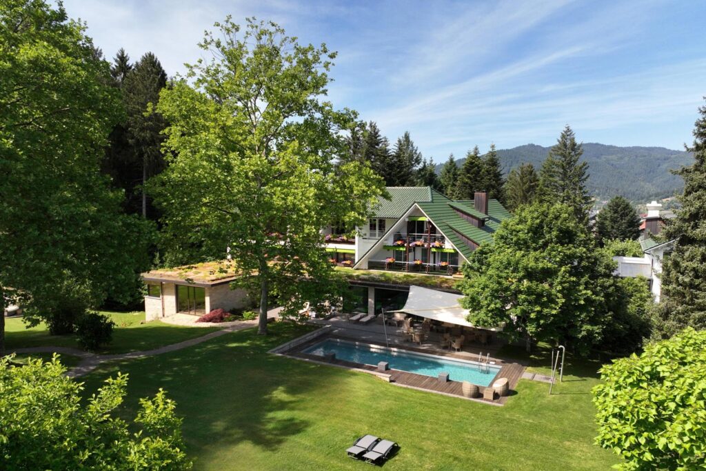 Zwischen hohen Bäumen und Bergen steht ein das Waldhotel Grüner Baum mit einem grünen Dach und Balkonen. Im weitläufigen Garten gibt es einen Swimmingpool, Liegestühle und einen gepflegten Rasen unter blauem Himmel mit dem Schwarzwald im Hintergrund.