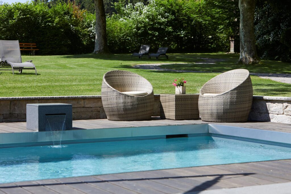 Zwei Korbstühle und ein kleiner Tisch mit einer Topfpflanze stehen an einem Pool auf einer Holzterrasse mit einer Steinmauer, üppigem grünen Rasen, Bäumen und Liegestühlen im Hintergrund.