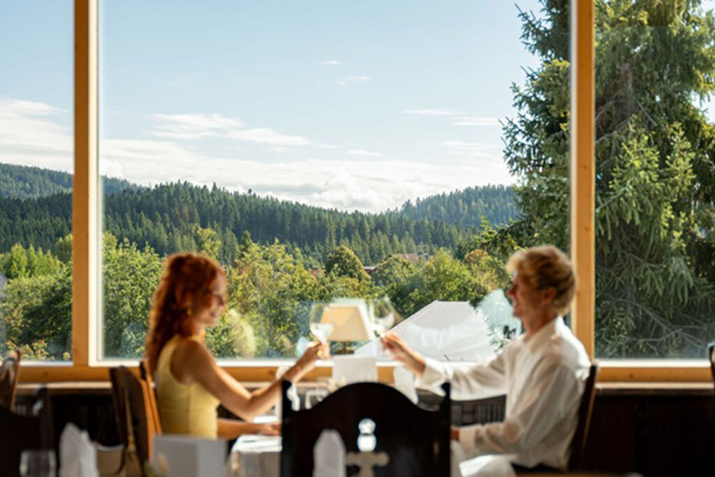 Zwei Personen sitzen an einem Tisch in einem Restaurant an einem großen Fenster und stoßen mit Weingläsern an. Draußen bietet sich ein malerischer Blick auf einen üppigen grünen Wald und Hügel unter einem hellen, klaren Himmel.