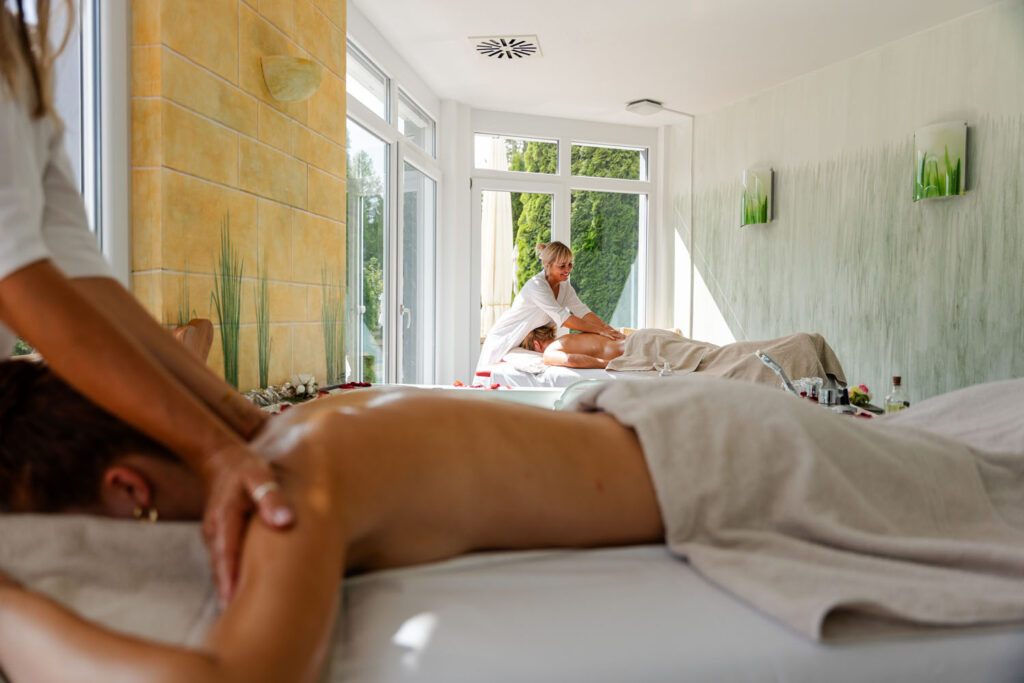 Zwei Personen werden in einem hellen, sonnendurchfluteten Wellness-Raum mit großen Fenstern massiert. Beide liegen auf Massagetischen, die mit weißen Handtüchern bedeckt sind, während die Therapeuten an ihren Rücken arbeiten. Die Atmosphäre wirkt ruhig und entspannend.