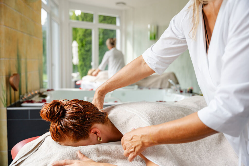 Eine Frau erhält eine Schultermassage, während sie mit dem Gesicht nach unten liegt, bedeckt mit einem Handtuch, in einem hellen, modernen Spa mit großen Fenstern und einem anderen Kunden im Hintergrund.