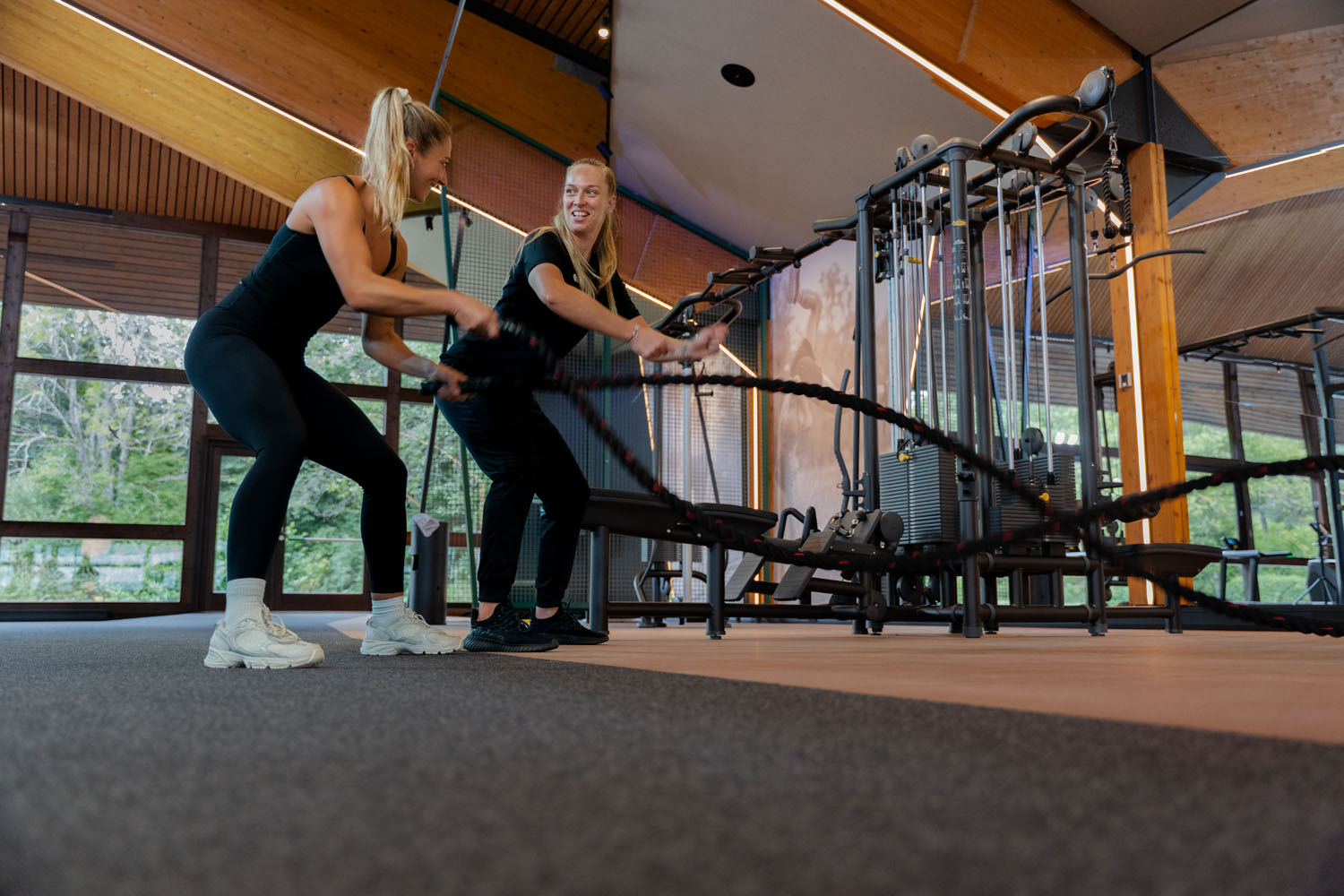 Zwei Frauen in Sportkleidung trainieren gemeinsam mit Kampfseilen in einem geräumigen, modernen Fitnessstudio mit großen Fenstern und Holzdecken.