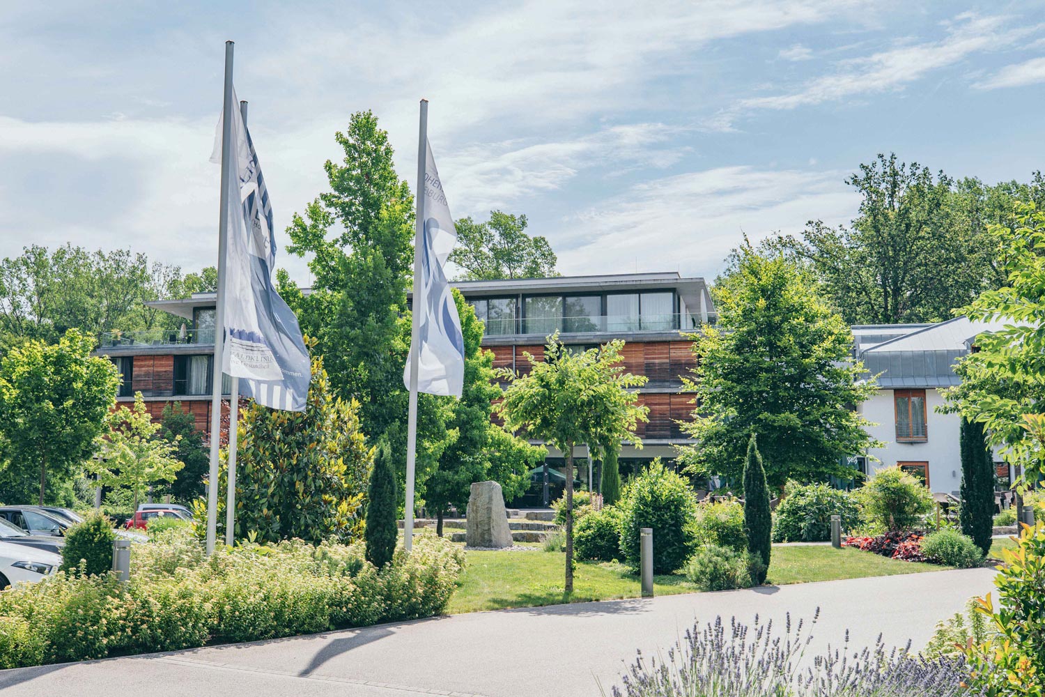 Ein modernes Gebäude mit großen Fenstern ist von üppig grünen Bäumen und angelegten Gärten umgeben. Zwei weiße Fahnen stehen auf Stangen im Vordergrund unter einem teilweise bewölkten Himmel.