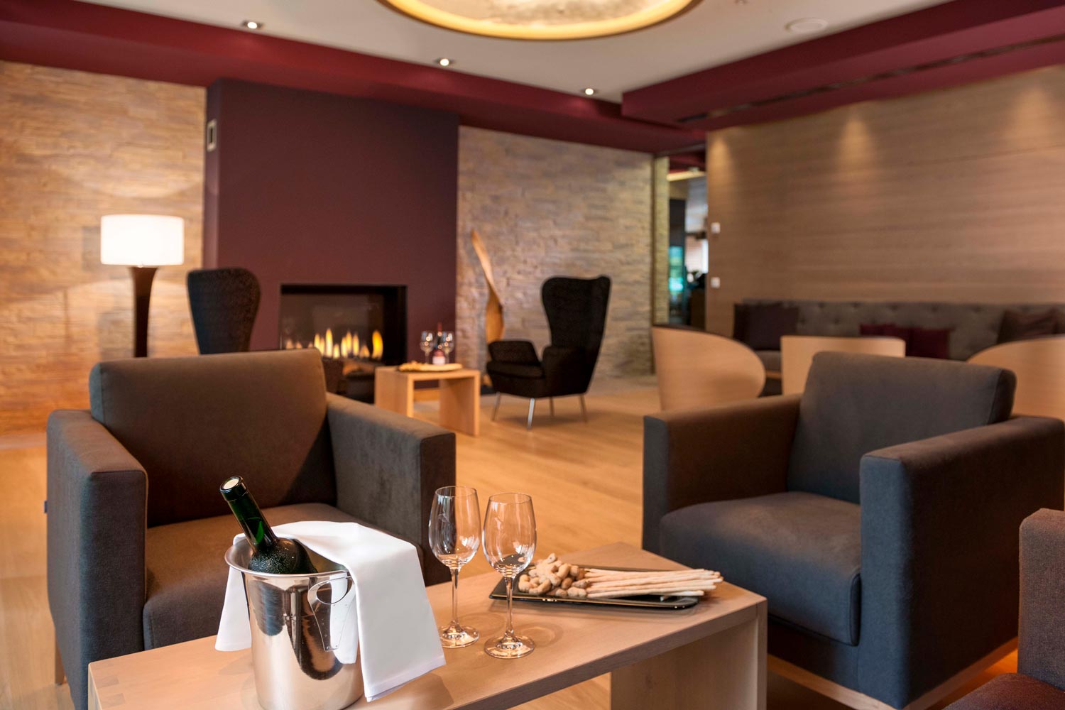 Eine moderne Lounge mit gemütlichen Sesseln, einem hölzernen Couchtisch mit Sekt, Gläsern und Snacks und einem beleuchteten Kamin im Hintergrund, der eine warme und einladende Atmosphäre schafft.
