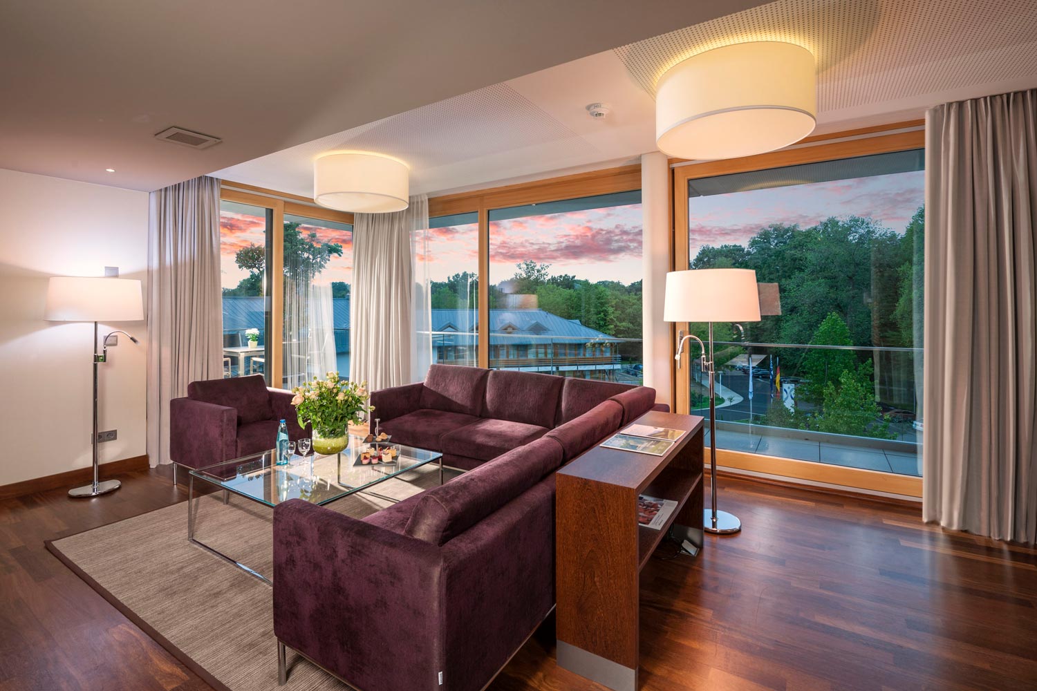 Ein modernes Wohnzimmer mit dunkelvioletten Sofas, einem Couchtisch aus Glas, großen Fenstern, Stehlampen und einem Blick auf Bäume und einen farbenfrohen Sonnenuntergang draußen. Der Raum ist mit Holzböden und farblich neutralen Vorhängen ausgestattet.