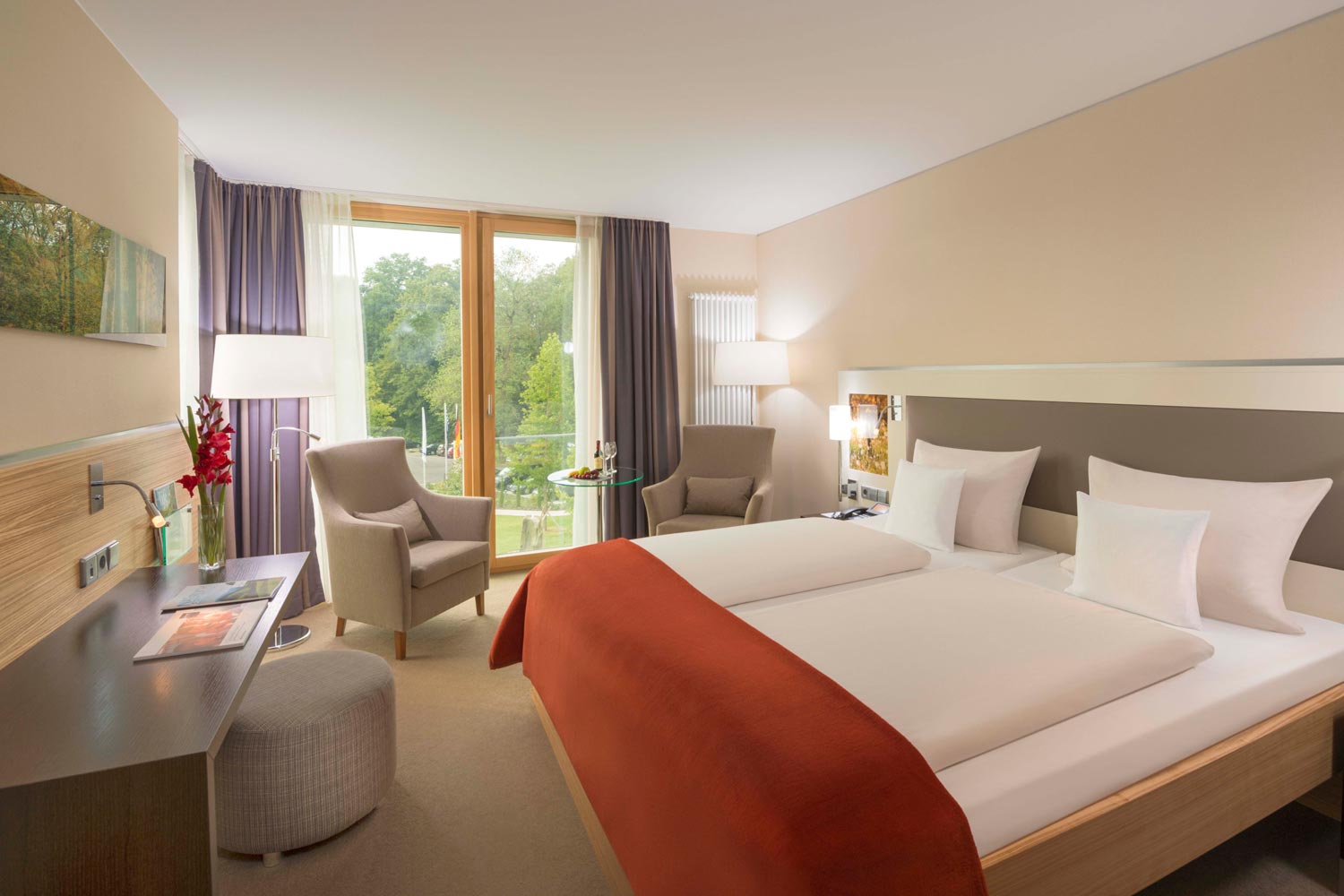 Ein modernes Hotelzimmer mit einem Doppelbett, einer orangefarbenen Decke, einem Schreibtisch, zwei Stühlen, einem kleinen Tisch und großen Fenstern mit Blick auf die grüne Umgebung. Das Zimmer ist gut beleuchtet und in neutralen Tönen gehalten.