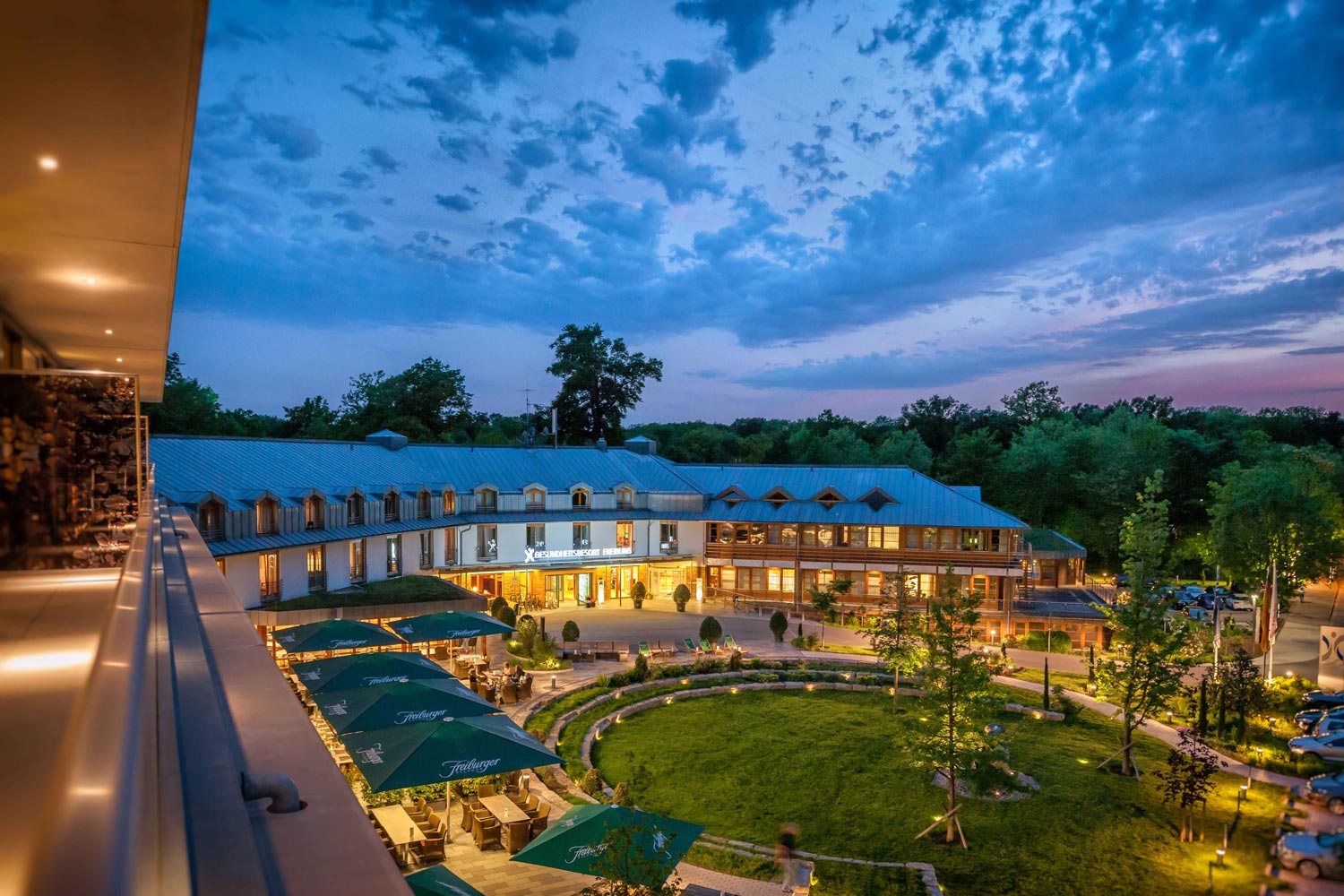 Ein großes, modernes Hotel mit Sitzgelegenheiten im Freien und grünen Rasenflächen wird in der Abenddämmerung beleuchtet, umgeben von Bäumen unter einem dramatischen blauen und violetten Himmel mit vereinzelten Wolken.