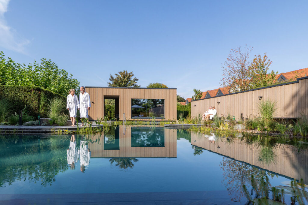 Zwei Personen in weißen Gewändern spazieren neben einem modernen Außenpool mit klarem Wasser, umgeben von Grün und einem Holzzaun; eine weitere Person in einem Gewand sitzt im Hintergrund unter einem blauen Himmel.
