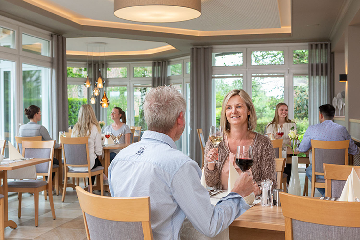 Ein lächelndes Paar sitzt an einem Restauranttisch und stößt mit Weingläsern an. Im Hintergrund sind andere Gäste zu sehen. Der Raum ist hell, mit großen Fenstern und moderner Einrichtung.