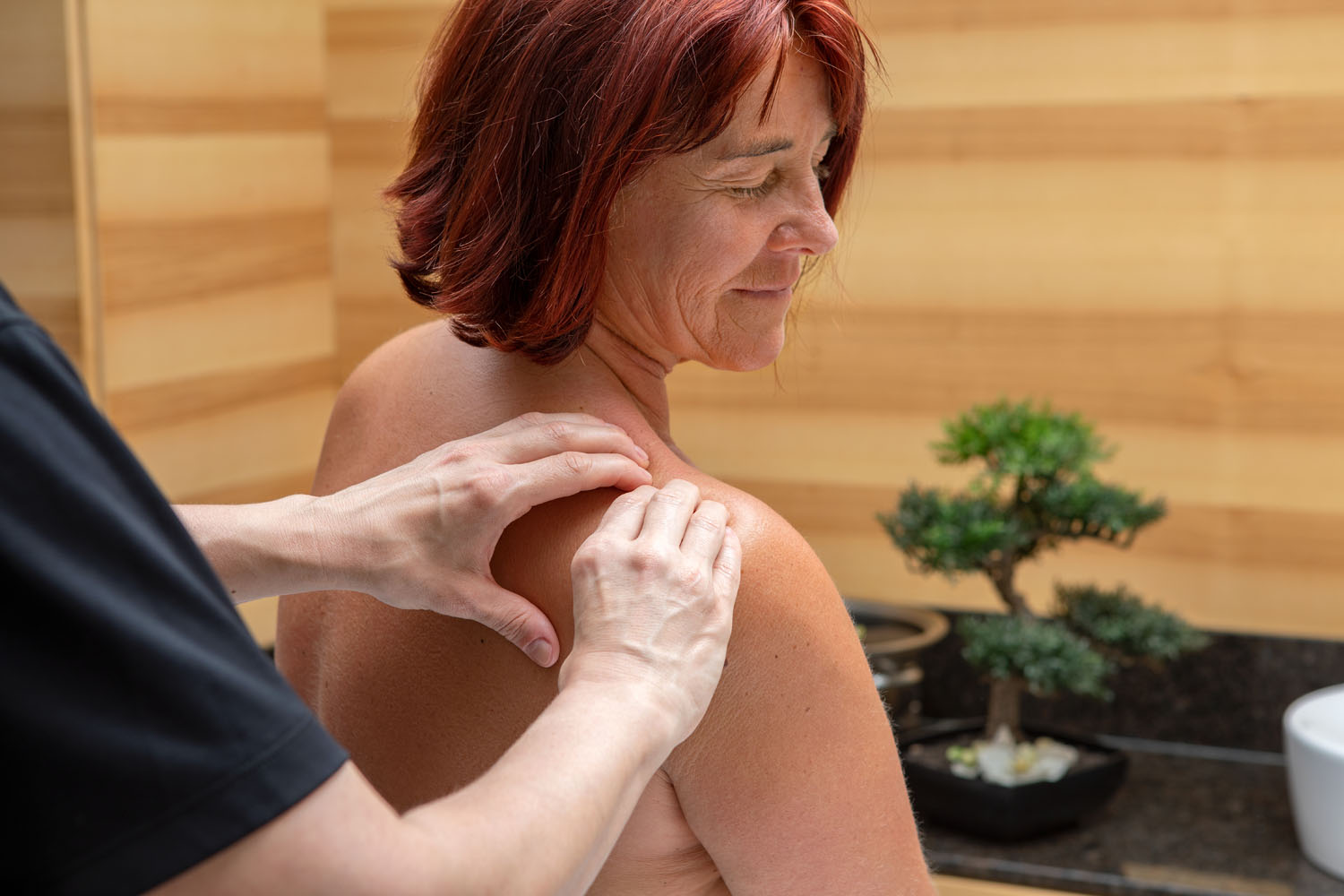 Eine Frau mit kurzen roten Haaren wird in einem ruhigen Raum mit Holzwänden und einem kleinen Bonsai-Baum im Hintergrund von einer Person in einem schwarzen Hemd an den Schultern massiert.